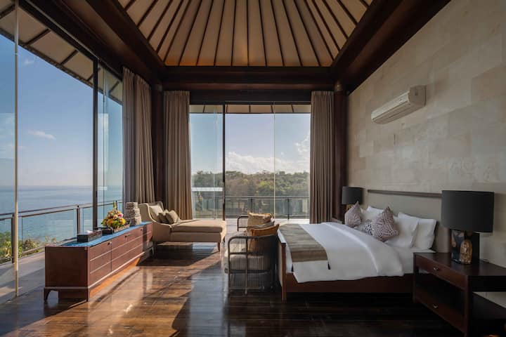A spacious bedroom with large windows offering stunning views of the ocean and dramatic cliffs. The room is filled with natural light, featuring a king-sized bed, minimalistic decor, and soft, neutral tones.