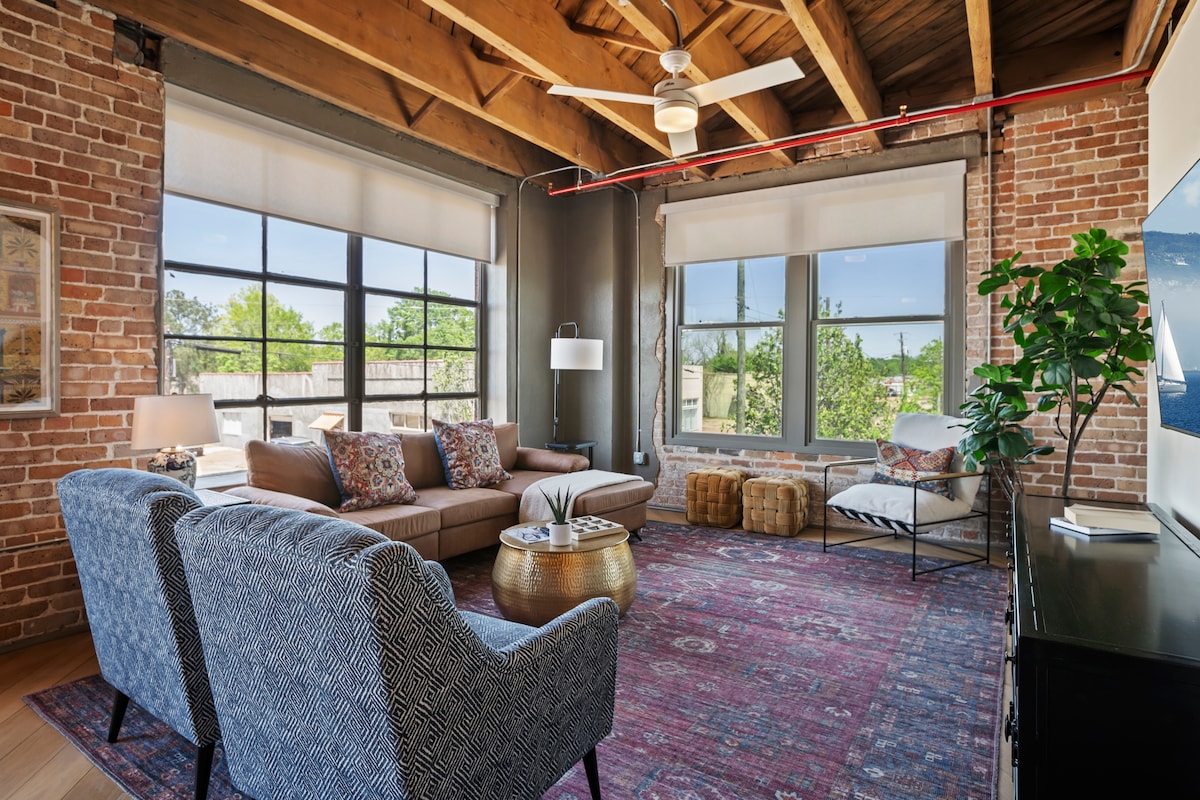The living area features exposed brick walls and wooden beams, providing a loft-style ambiance. A comfortable sofa, accent chairs, and a decorative area rug contribute to a cozy atmosphere. Large windows allow for natural light, overlooking greenery and the outdoors.