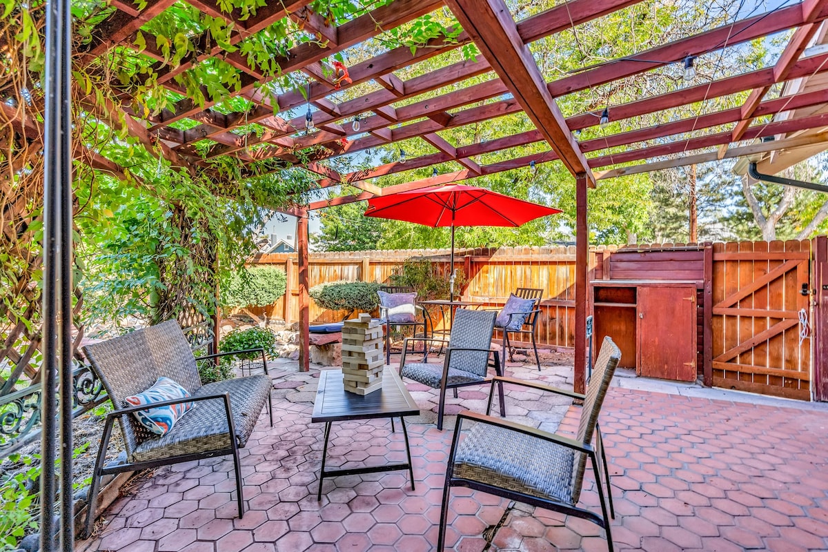 An outdoor space is presented with a shaded pergola featuring a vibrant red umbrella over a small table. Arranged around the table are multiple chairs, with low greenery and a wooden barrier enclosing the area. A grill is positioned to one side, and a game set is visible.