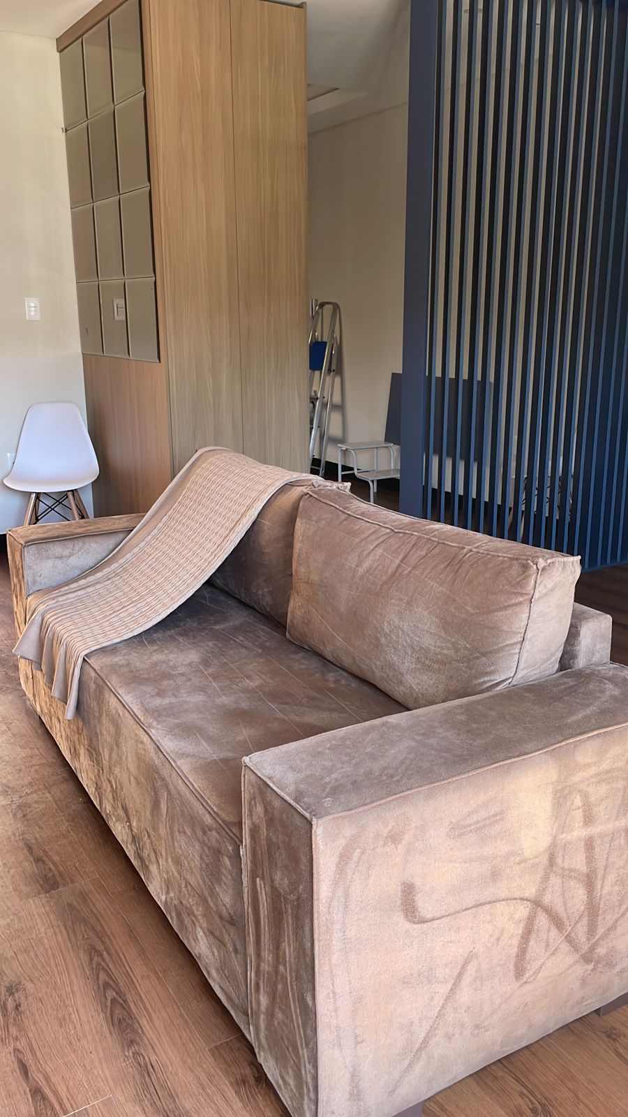 A modern, comfortable sofa with soft upholstery is placed in a well-lit room. A textured throw is draped casually over one side. The background features wooden cabinetry and sleek, vertical panels adding depth to the space.