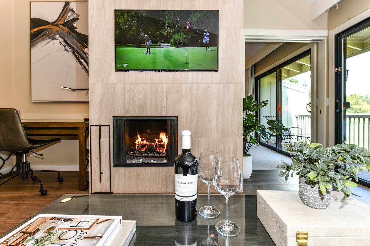A modern living space features a sleek fireplace surrounded by neutral-toned stonework. A bottle of wine sits on a glass table, accompanied by two glasses ready for use. The room’s design is complemented by a large TV mounted above the fireplace, showcasing a golf scene.