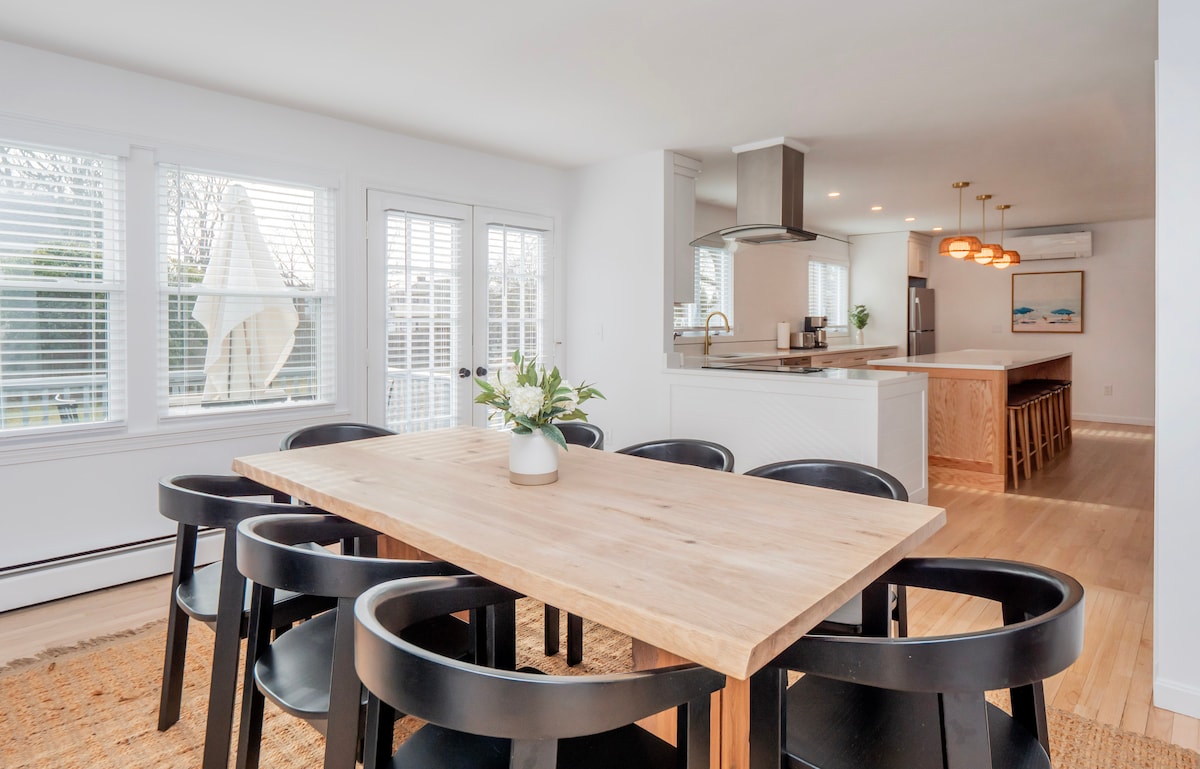 A dining area is highlighted by a wooden table surrounded by black chairs. Large windows allow natural light to fill the space, and the nearby kitchen features a modern layout with bar seating and pendant lighting. A neutral color palette contributes to the room's fresh ambiance.