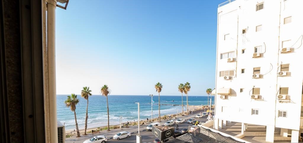 A clear view of the coastline is presented, featuring gently rolling waves and palm trees lining the beach. The busy promenade is visible, with vehicles driving along the road. The scene captures the vibrant atmosphere of the beachfront area under a bright blue sky.