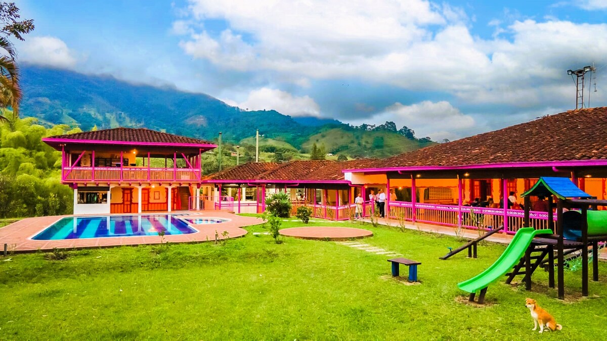 A vibrant outdoor view of Kiara Ecohotel showcases colorful buildings set against a mountainous backdrop. A large pool is surrounded by lush green grass, while a children’s playground features slides and swings. The tranquil atmosphere is complemented by soft clouds overhead.