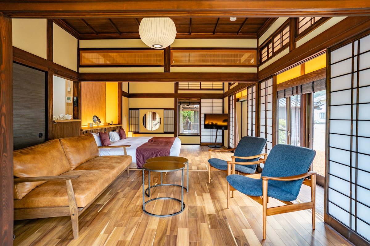 A spacious living area features large windows that allow natural light to fill the room. A comfortable sofa and two armchairs are arranged around a circular table, with wooden flooring and traditional sliding doors creating a harmonious atmosphere. A bed area is visible in the background.