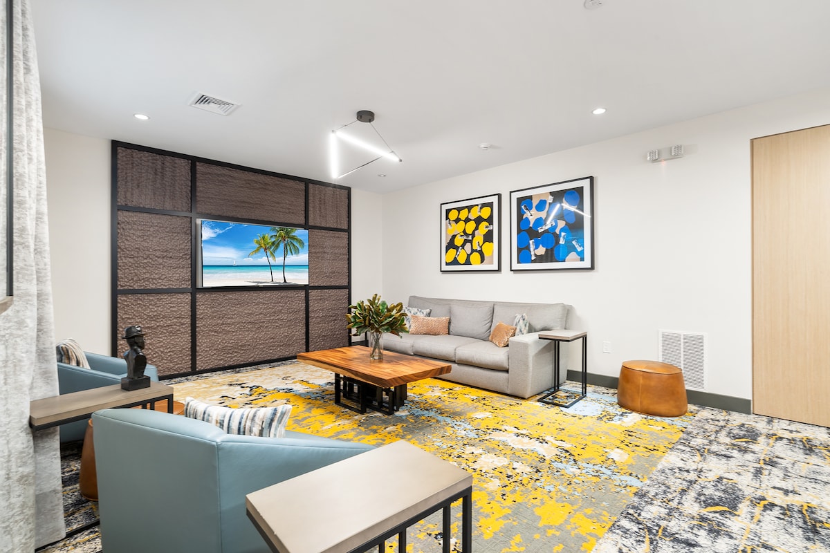 The living room features a convertible sofa, two blue armchairs, and a coffee table. A large window displays a beach view with palm trees. Colorful artwork adorns the walls above the sofa, and a patterned rug adds texture to the space.