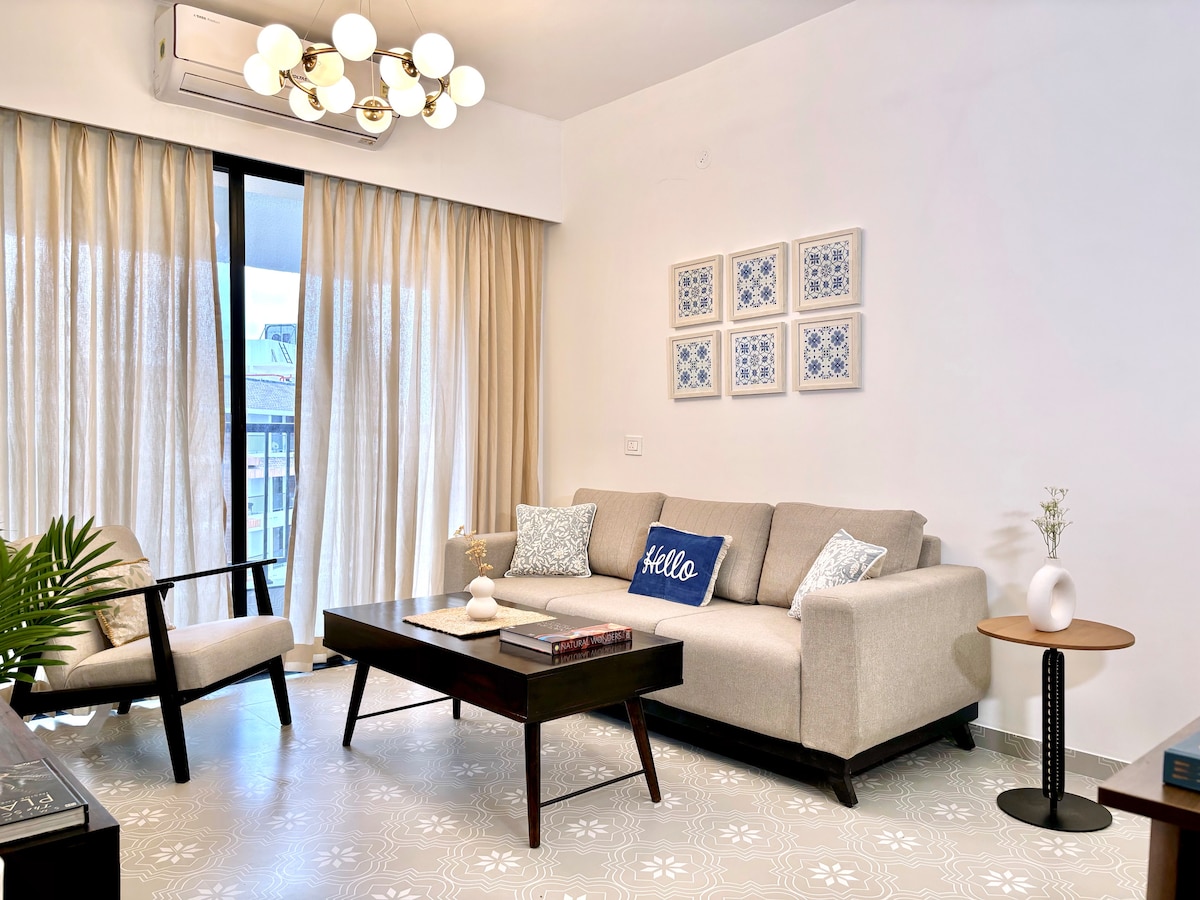 A well-lit living area features a comfortable, beige sofa paired with accent pillows, complemented by an armchair. A coffee table sits centrally, resting on a patterned floor. Large glass doors invite natural light, while sheer curtains softly frame the view.