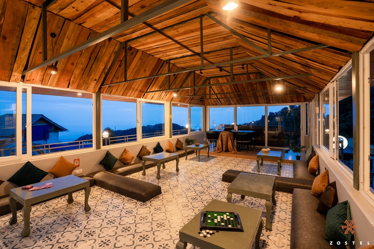 A spacious communal area features a wooden ceiling with exposed beams, offering a relaxed atmosphere. Floor cushions are arranged around low tables, while large windows frame views of the landscape, inviting natural light into the space. A patterned tile floor adds character to the room.