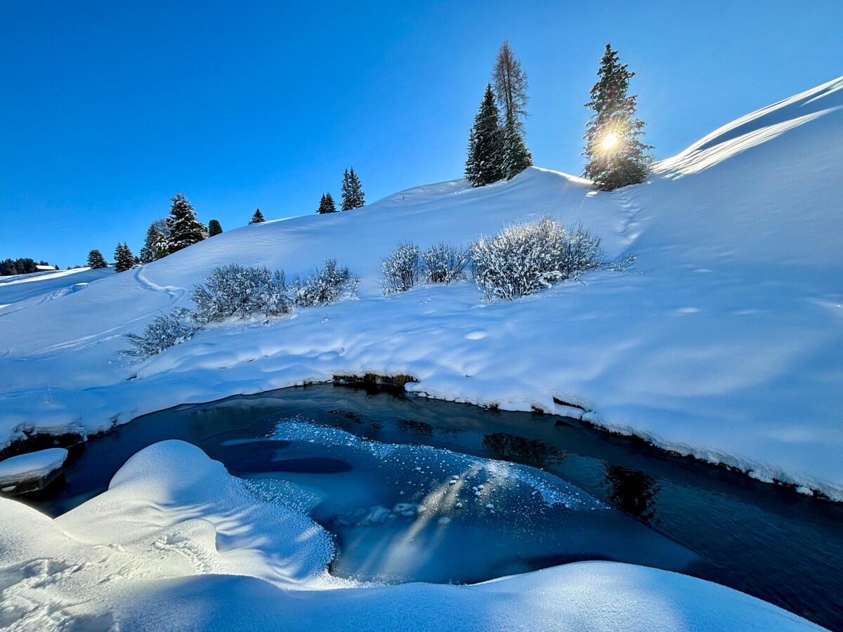 A serene winter landscape is depicted, featuring a gently flowing stream bordered by soft, snowy mounds. Sparse bushes are visible, partially covered in snow. Sunlight illuminates a distant hill and trees, casting faint shadows on the pristine white snow.