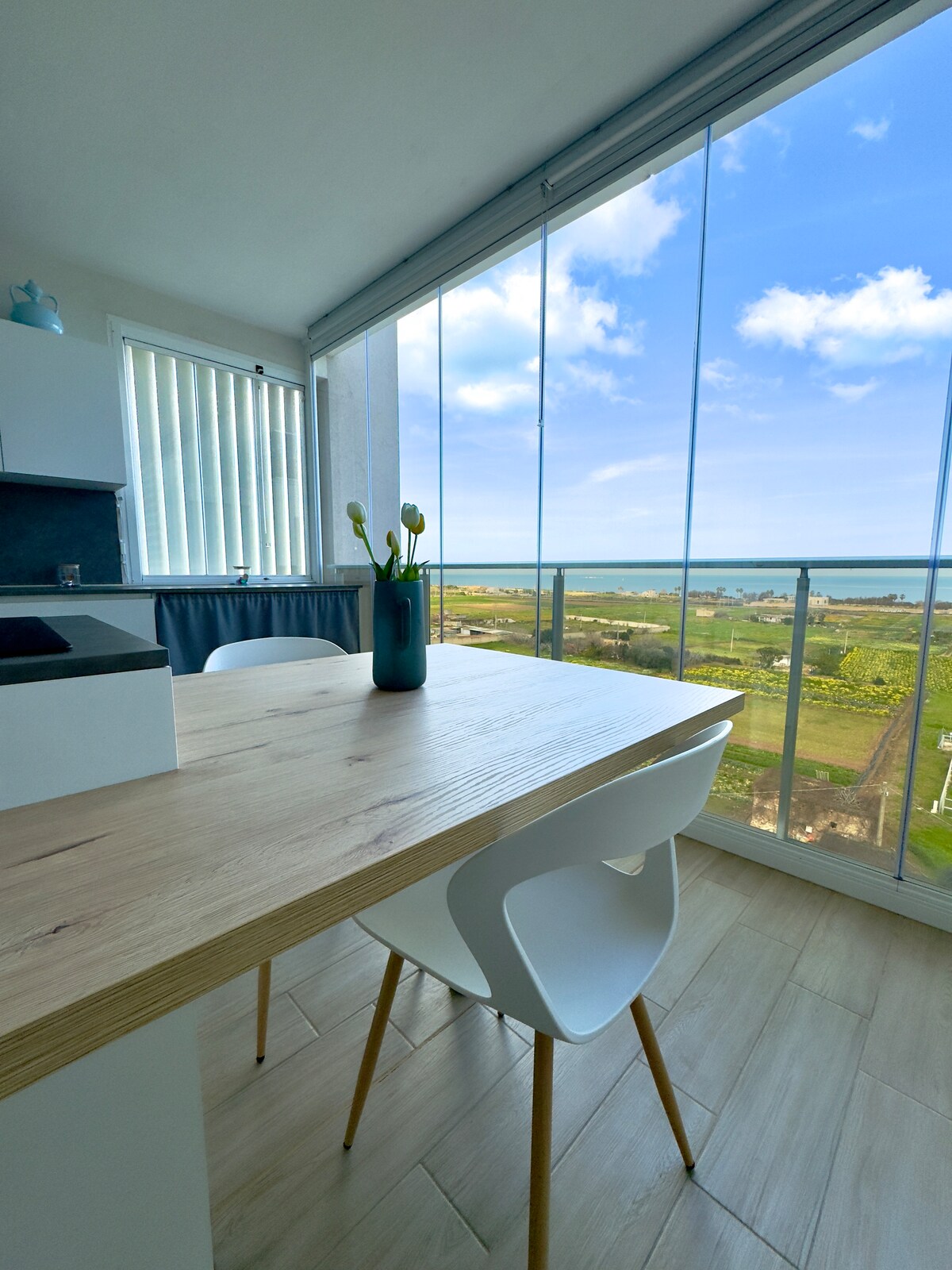 A dining table with modern white chairs is positioned near large glass windows, offering expansive views of the sea and surrounding landscape. Natural light floods the space, creating a bright and open atmosphere. A small vase with flowers adds a touch of color to the table.