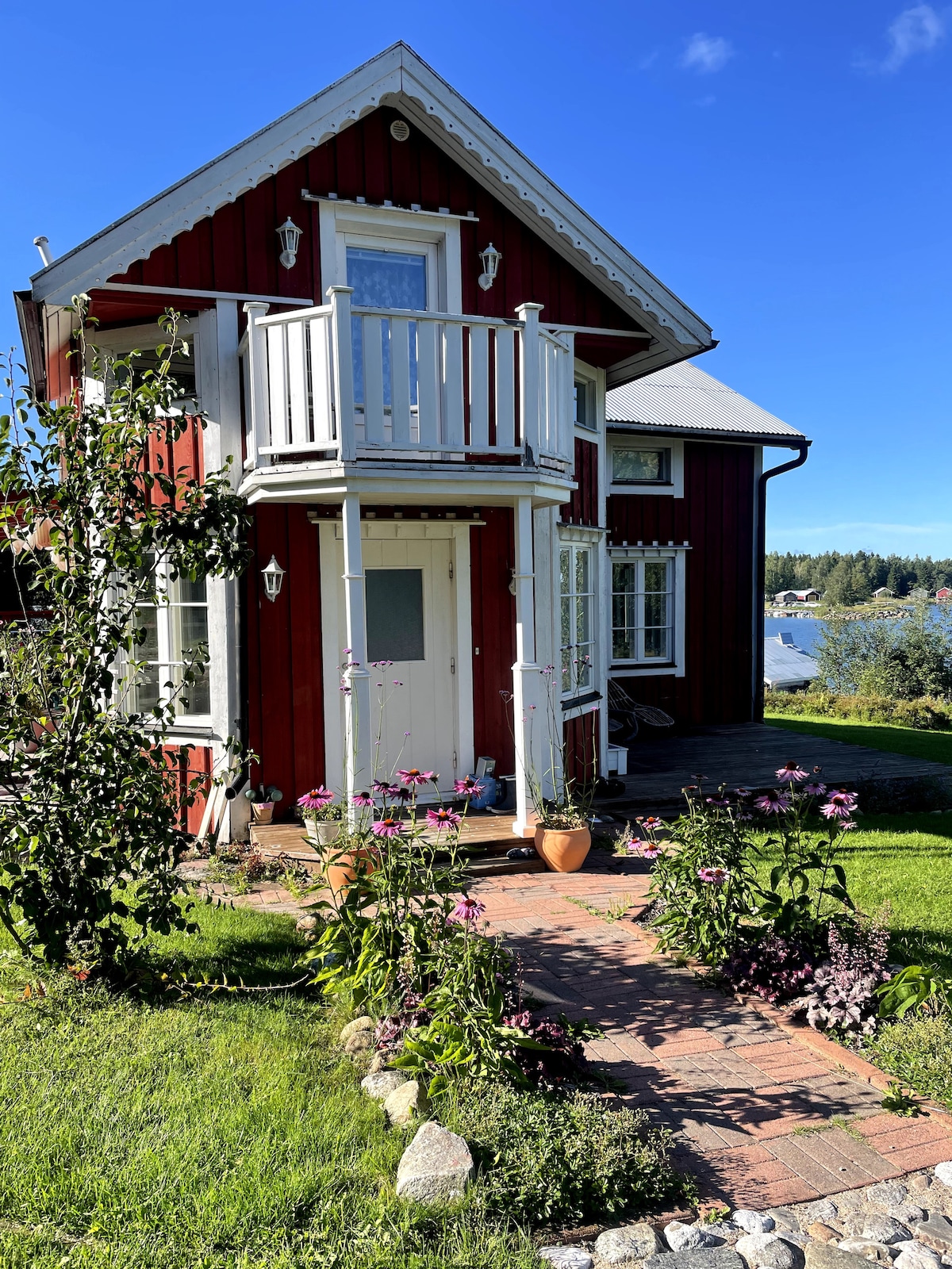 The exterior of a red wooden house is displayed, featuring a welcoming entrance and a small balcony above. Lush green grass surrounds the property, with a flower-lined pathway leading to the door. Boathouses can be seen in the background, against a clear blue sky.