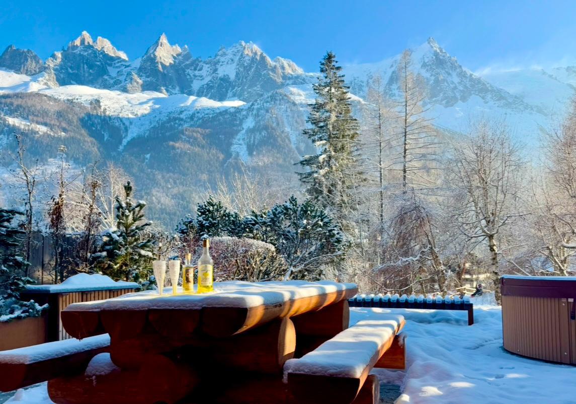 A wooden table and benches are covered in a light dusting of snow, positioned on a terrace. The backdrop features majestic snow-capped mountains, flanked by evergreen trees and bare winter branches. A clear blue sky adds brightness, enhancing the serene winter landscape.