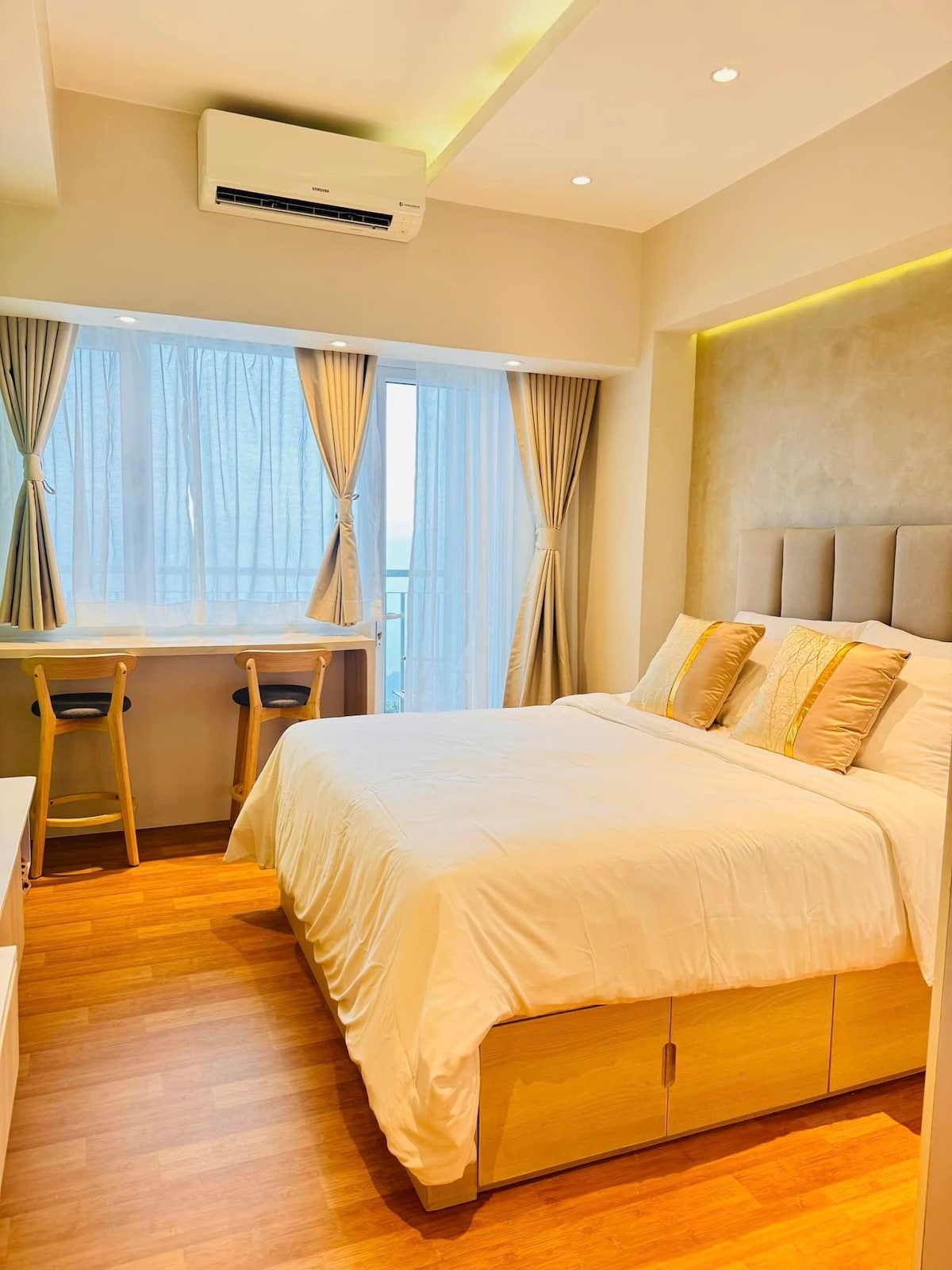 A bright bedroom is featured, featuring a spacious bed with a light-colored comforter and two accent pillows. A window is dressed with sheer curtains, allowing natural light to flow in. A small desk with two contemporary stools is positioned beside the window.