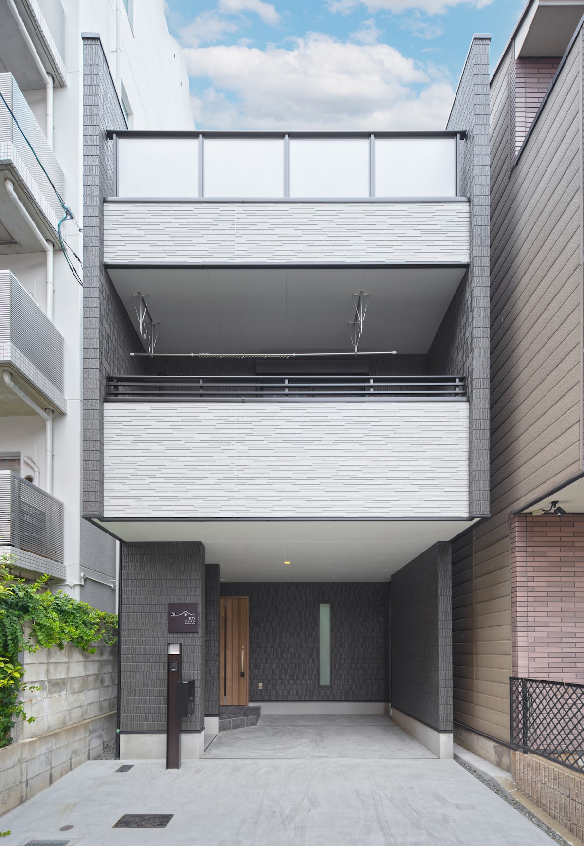 The exterior of a modern three-story home is displayed, featuring a minimalist design with a combination of textured gray and white materials. A balcony is visible on the second floor, framed by transparent panels. The entrance is marked by a sleek door and a subtle light fixture.