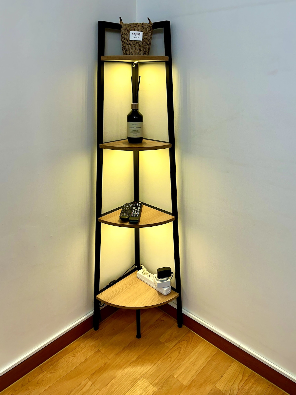 A corner shelf unit is showcased against a white wall, featuring four wooden shelves with an ambient light effect. Decorative items, including a wine bottle, a remote control, and a small plant, are placed on the shelves, enhancing the organized appearance.