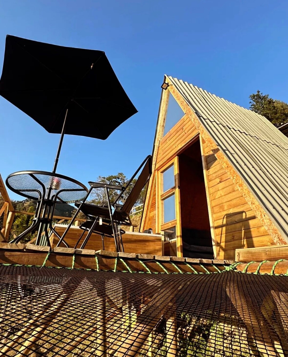 A charming A-frame structure is showcased against a clear blue sky. The outdoor seating area features a round table and two chairs, complemented by a large, dark umbrella providing shade. Warm wooden accents highlight the building's design, inviting relaxation in a natural setting.