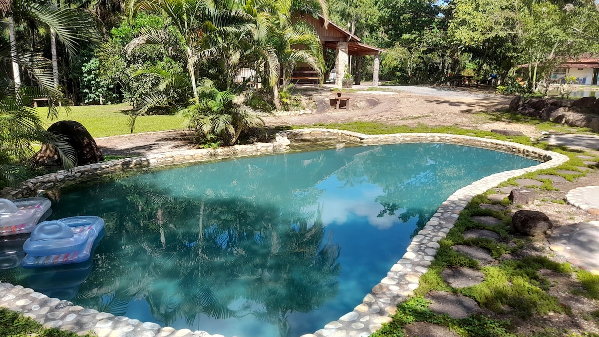 A natural swimming pool features clear water, bordered by smooth stones and surrounded by lush greenery. Floatation devices are present on the water's surface. A pathway of stones leads toward the house, with a spacious lawn visible in the background.