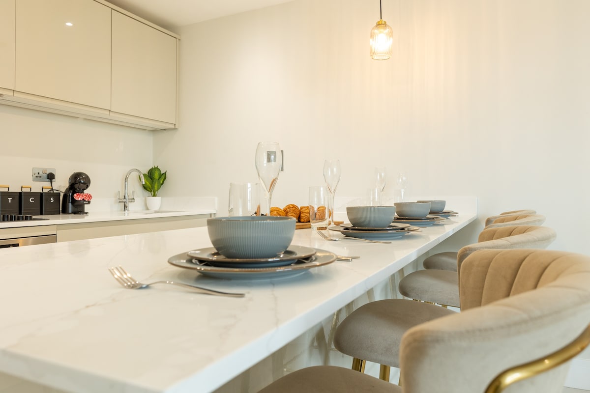 A sleek dining area features a polished marble counter with stylish dinnerware arranged for six. Plush chairs are positioned around the counter, while a decorative plant and pendant lighting add a touch of elegance. Soft, neutral tones create a welcoming atmosphere.