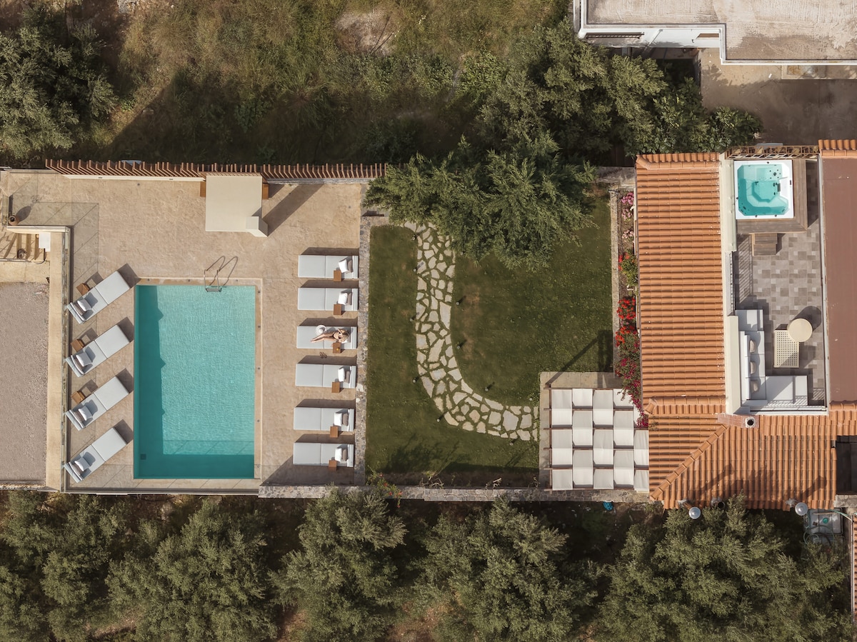 An aerial view of the villa's outdoor area showcases a private swimming pool surrounded by sun loungers. A well-maintained garden features a stone pathway leading to a relaxation area with comfortable seating, while a jacuzzi is visible on the terrace adjacent to the main house.