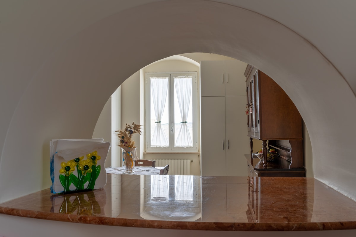 The kitchen area features an arched opening that frames a view of two windows adorned with sheer white curtains. A wooden cabinet is visible alongside a countertop with a marble finish. A decorative item with flowers sits on the counter, adding a touch of character.