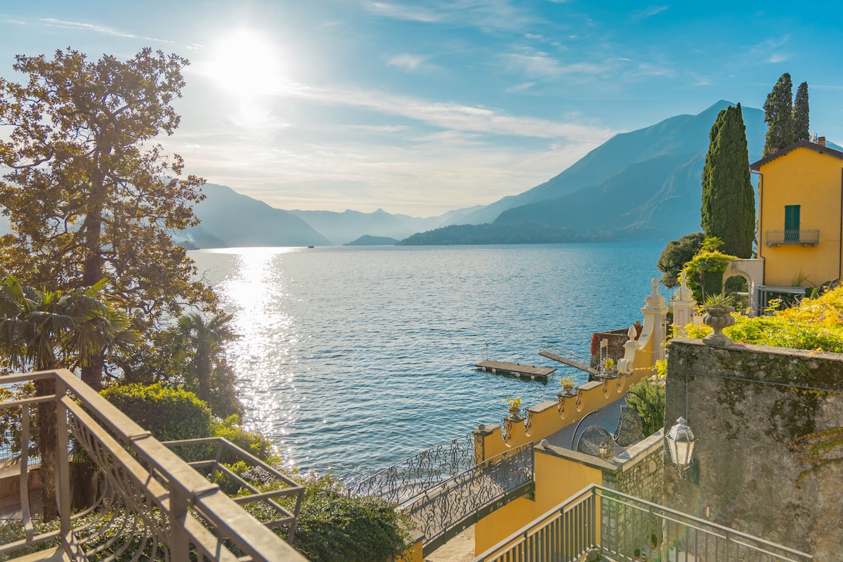 A serene view of the lake is captured, showcasing sparkling water reflecting sunlight. Surrounding trees and hills add a natural backdrop, while distant mountains frame the scene. A waterfront area can be seen, enhancing the tranquil atmosphere of this picturesque location.