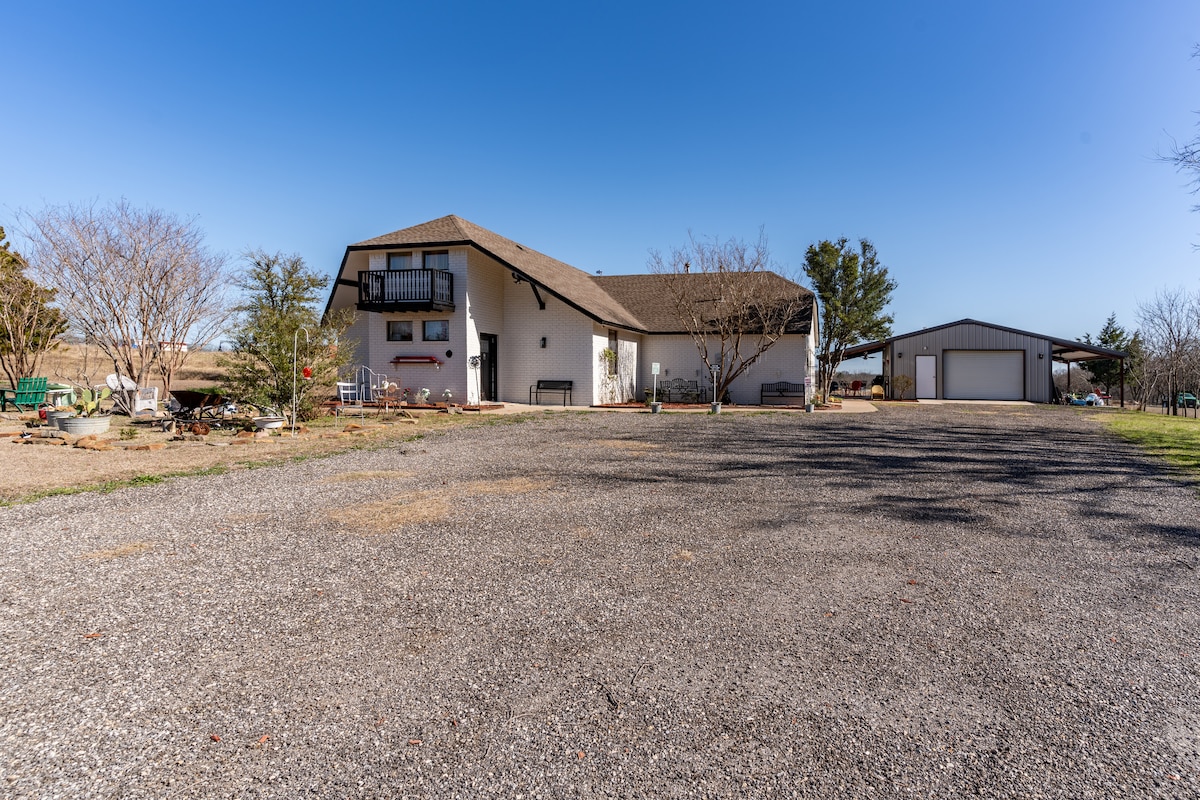 A spacious white structure with a sloped roof is set in a gravel driveway. Surrounding landscaping includes various trees and shrubs, with a separate building visible in the background. The setting offers an open, accessible outdoor space under a clear blue sky.