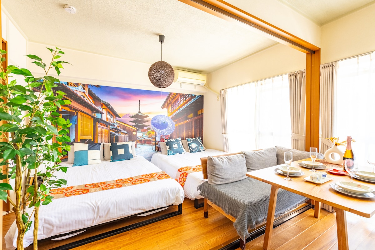 A spacious room is furnished with two double-sized beds adorned with patterned throws. A dining area with a wooden table and comfortable seating is visible. A large mural depicting a traditional Japanese street scene enhances the atmosphere, complemented by natural light from windows with sheer curtains.