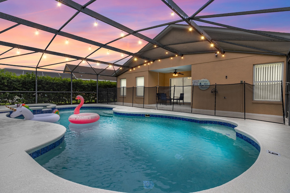 A large, inviting pool area is enclosed by a protective screen, featuring a gently shaped pool surrounded by a spacious deck. Colorful inflatable toys are present, and an outdoor dining set is positioned nearby, all under twinkling string lights against a serene sunset sky.