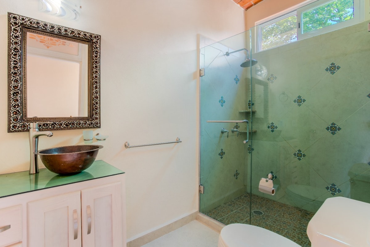 A modern bathroom features a walk-in glass shower, equipped with a rain showerhead. A stylish circular sink is set atop a green countertop, complemented by a decorative mirror. Natural light filters in through a window, and subtle tile accents add visual interest to the space.