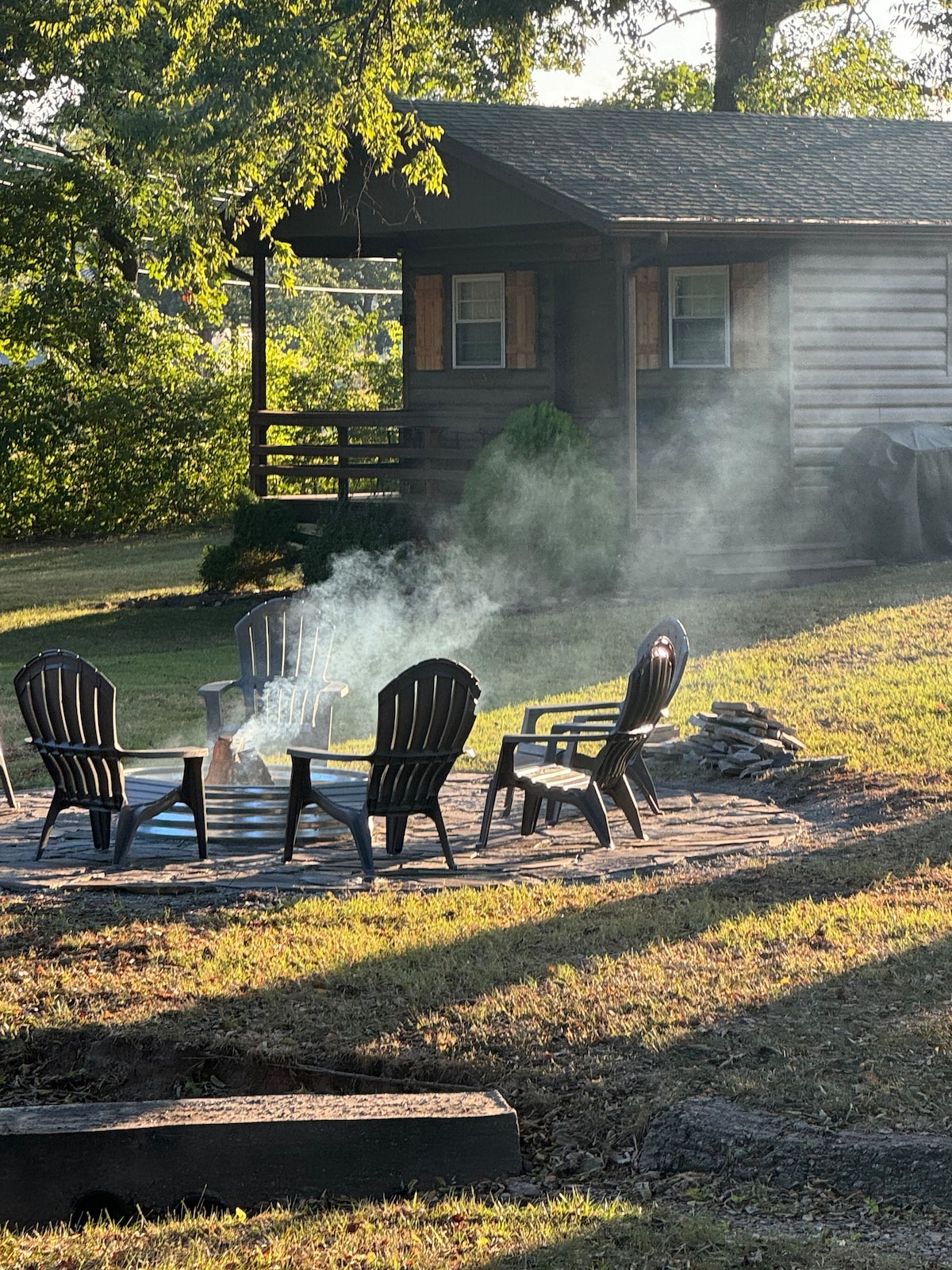 A circular stone firepit is surrounded by six black Adirondack chairs. Wisps of smoke rise into the air, hinting at a recently enjoyed fire. In the background, a wooden cabin is partially shaded by trees, creating a serene outdoor setting.