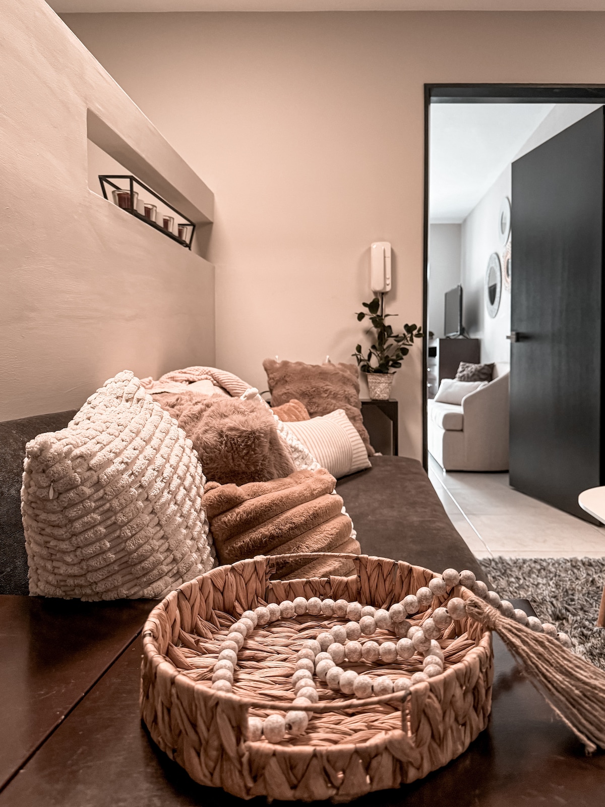 A cozy living area is presented with a dark sofa adorned with textured cushions in warm tones. A woven basket, filled with decorative elements, rests on a wooden table, enhancing the inviting atmosphere. A view into the adjoining room is visible through an open doorway.