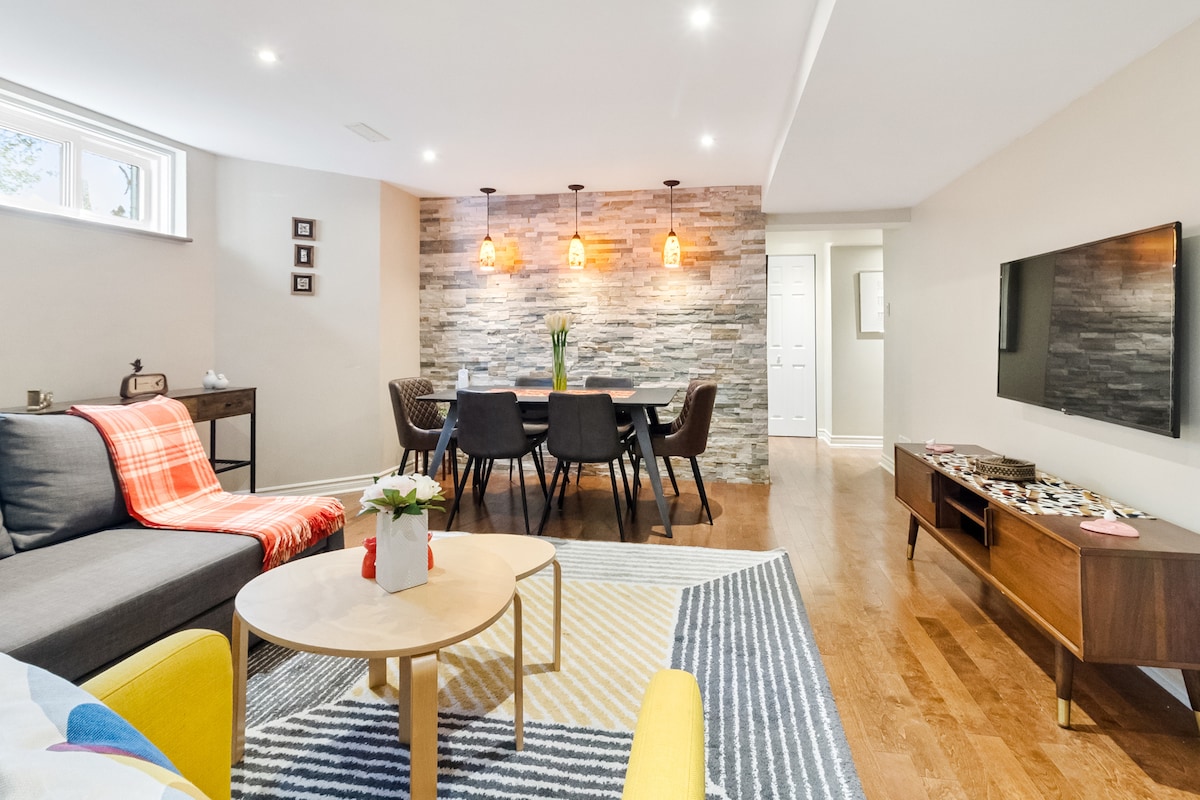 A comfortable living area is presented, featuring a sofa with a colorful throw and an adjacent coffee table. A dining space with dark chairs and pendant lighting is visible in the background, set against a stone accent wall. A wall-mounted TV and wooden media console complete the space.