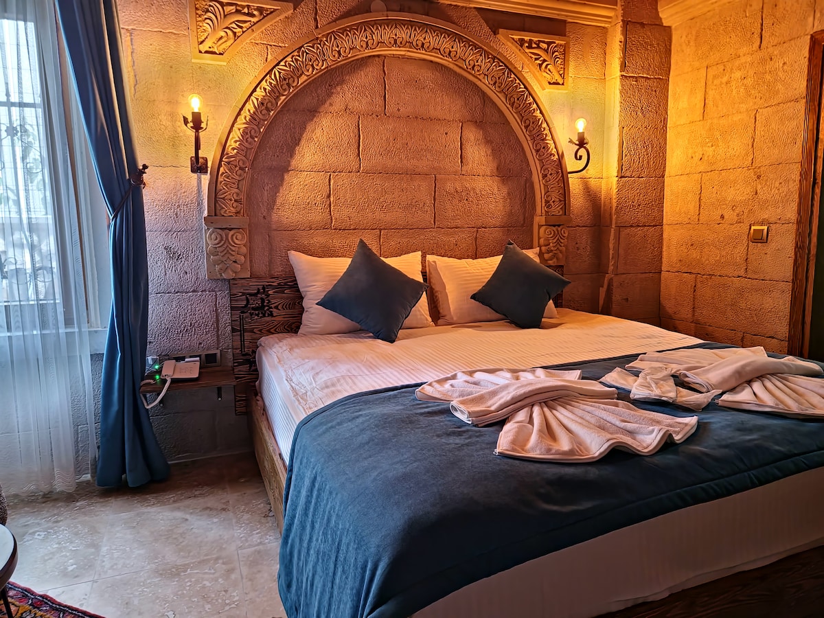 A spacious bed is positioned against a stone wall adorned with intricate carvings. Soft blue and white linens complement two decorative pillows. Warm lighting from wall sconces adds a cozy ambiance, while a window draped with a blue curtain allows natural light to filter in.