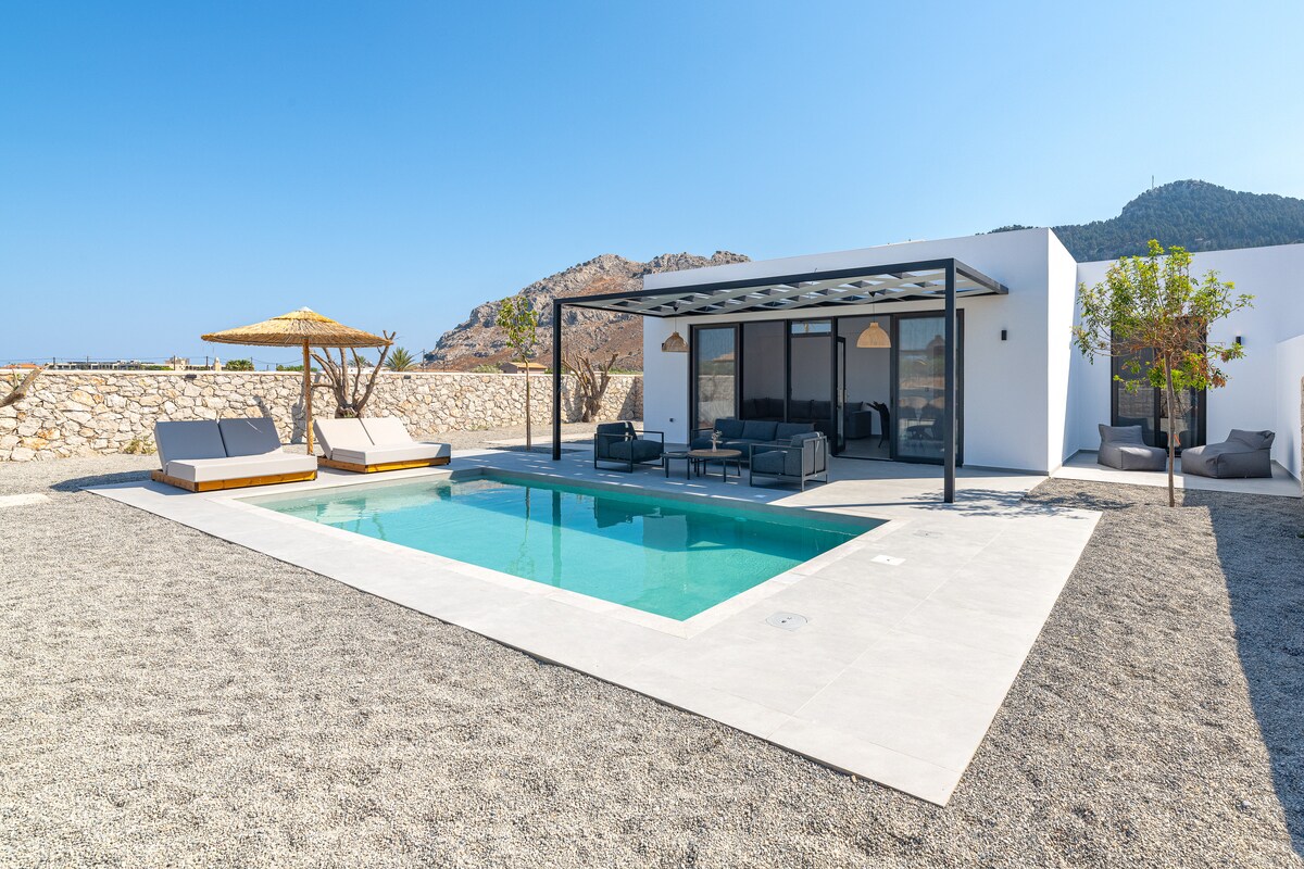 A private pool is surrounded by a spacious outdoor area featuring comfortable lounge chairs and shaded seating. The villa's modern architectural design is complemented by distant hillside views, while a sun umbrella provides additional shade for outdoor relaxation.