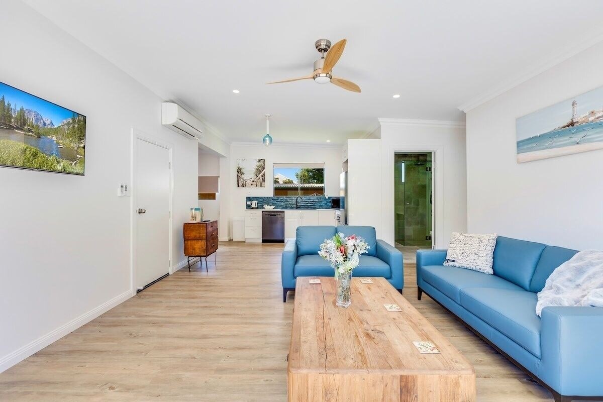 The open living area features modern blue sofas positioned around a wooden coffee table. A ceiling fan hangs above, and natural light fills the space through large windows. A kitchen area with stainless steel appliances is visible in the background.