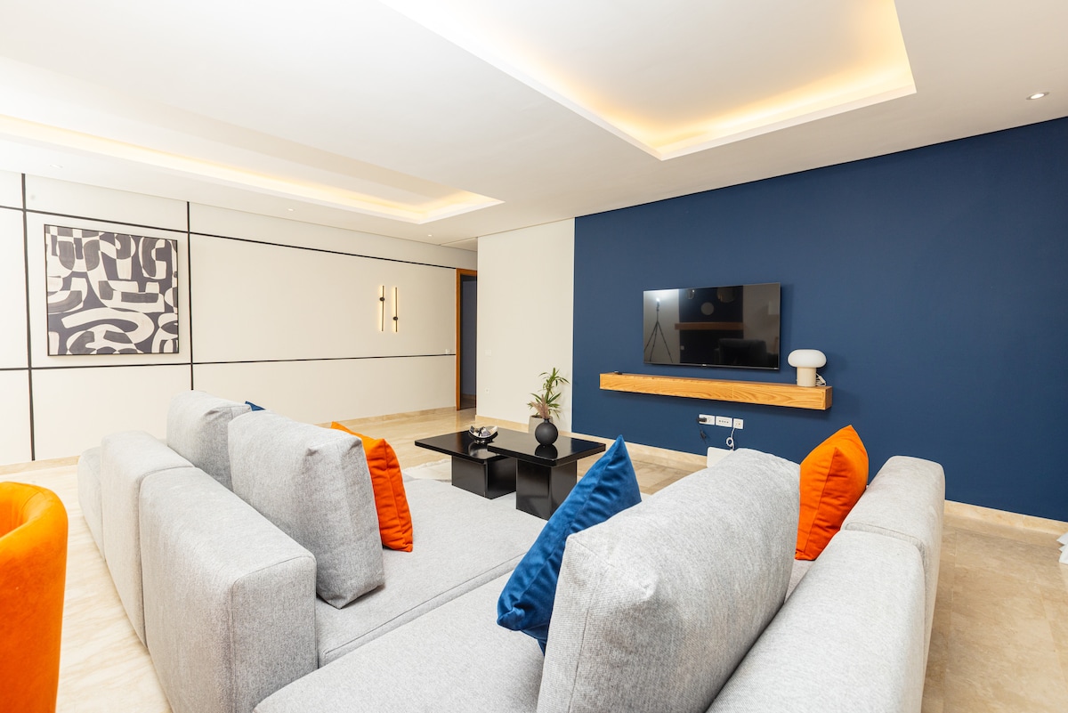 A spacious living area is highlighted by a large, gray sectional sofa adorned with orange and blue accent pillows. A sleek black coffee table is centered in front of a modern entertainment center featuring a flat-screen TV mounted on a contrasting blue wall.