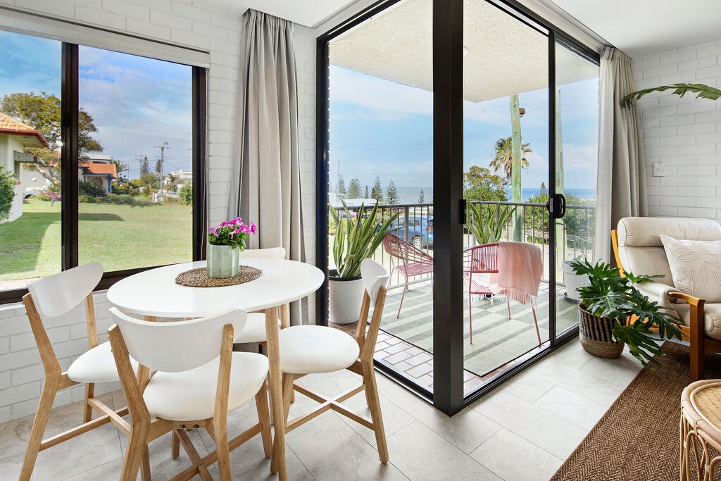 An inviting dining area features a round table surrounded by four light wooden chairs. Large glass doors open to a spacious balcony adorned with plants, overlooking lush greenery and distant ocean views. Natural light floods the room, complemented by subtle wall decor.