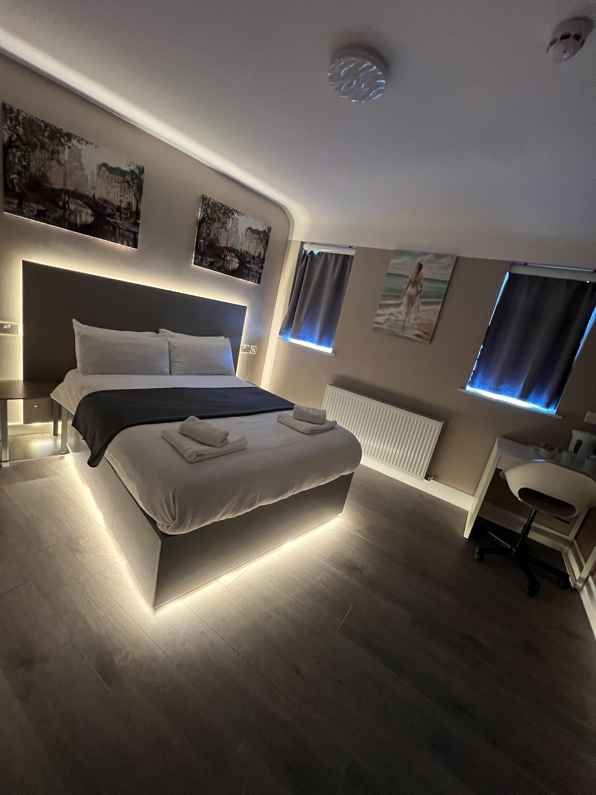 A modern room features a spacious bed with a lighted base, enhancing the contemporary design. Two windows allow natural light, softened by dark curtains. Artwork adorns the walls, and a desk and chair are positioned in one corner, providing a functional workspace.