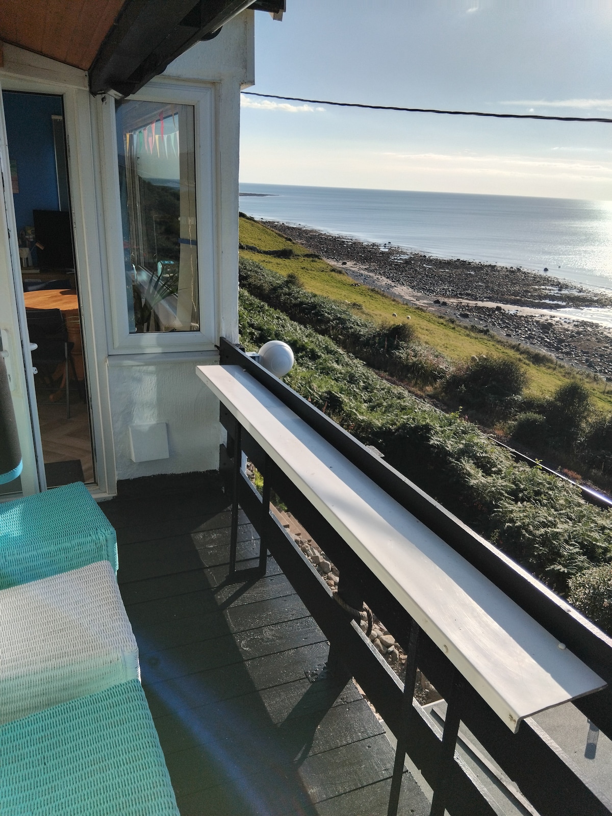 A balcony view features a railing overlooking a rocky shoreline and the tranquil sea. Sunlight reflects off the water, and a grassy hillside rises in the background. The scene is framed by the cottage's adjoining structure, offering an inviting spot to relax.