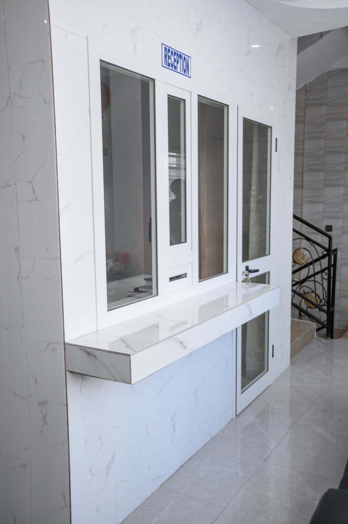 A sleek reception area is showcased with a glossy white surface and clean lines. The window arrangement features three panes, allowing for communication, while a sign above indicates 'RECEPTION.' The tile flooring reflects natural light, contributing to a bright and modern ambiance.