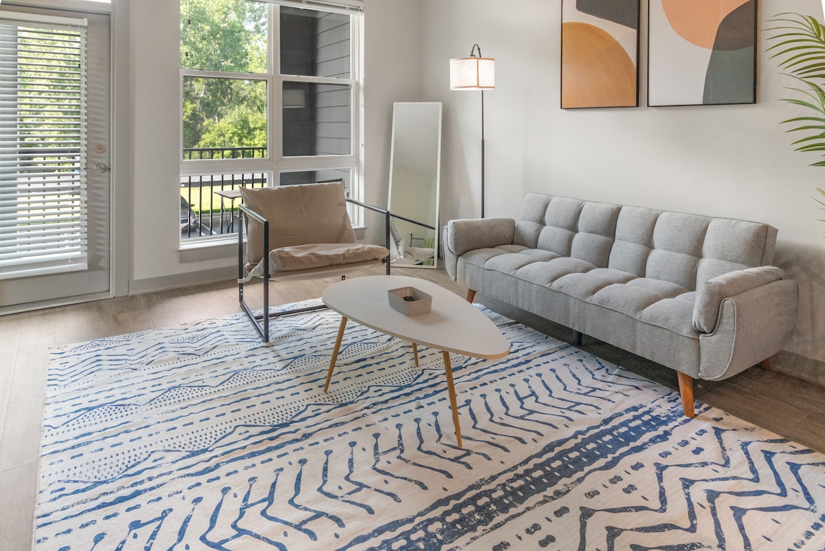 A modern living area showcases a gray sofa and a minimalist chair, complemented by an abstract artwork collection on the wall. A light rug with blue patterns lies beneath a coffee table, and natural light filters in through large windows, enhancing the open atmosphere.