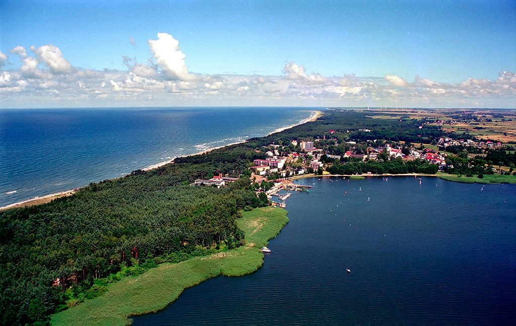An aerial view captures a coastal landscape where the blue sea meets a lush forest. The shoreline stretches into the distance, bordered by sandy beaches. A quaint town is visible near the water, with a mix of greenery and buildings contributing to a peaceful atmosphere.