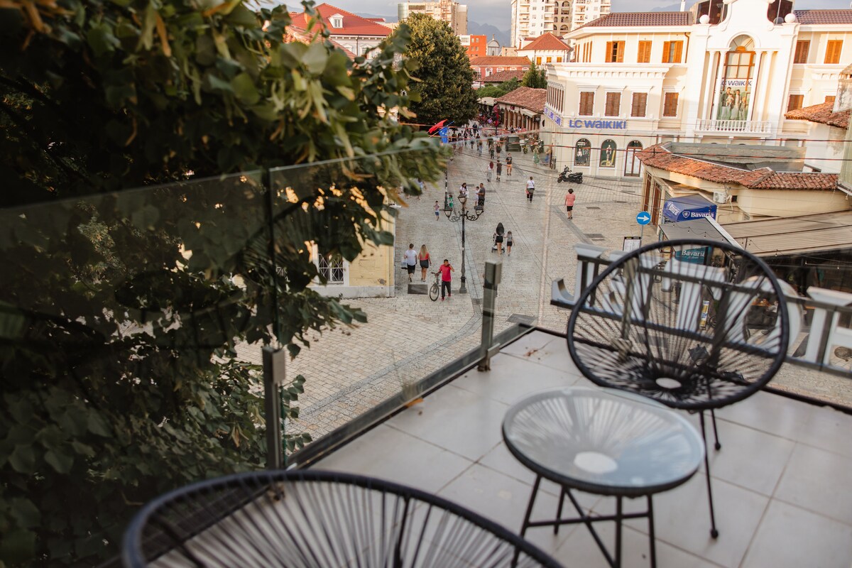 A spacious terrace features two round, black chairs positioned overlooking a lively pedestrian street. The scene captures people walking and cycling below, highlighting the vibrant atmosphere of the surrounding area. Lush trees frame the view against a backdrop of historic architecture.