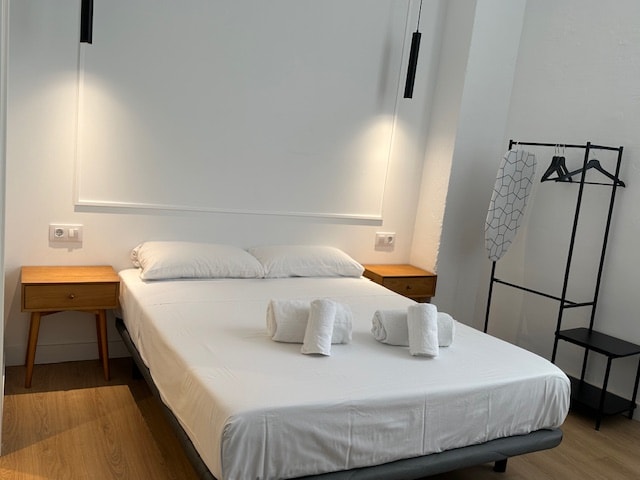 A bright sleeping area features a spacious double bed dressed in fresh white linens. Two bedside tables are positioned on either side, with wall-mounted light fixtures providing soft illumination. A clothing rack stands nearby, offering additional storage space.