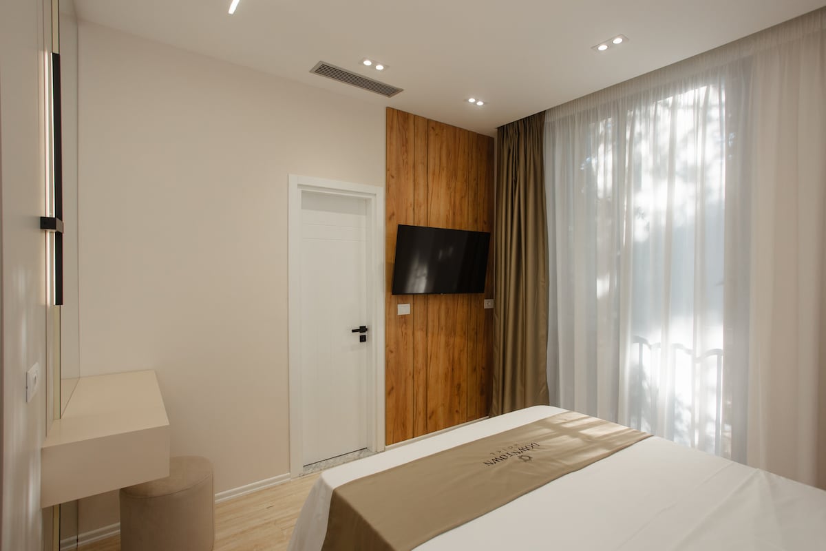 A modern guest room features a spacious bed with neutral-toned bedding. A wooden accent wall adds texture, complemented by a flat-screen TV mounted on the opposing wall. Natural light filters through sheer curtains, enhancing the serene ambiance. A minimalist desk and stool are positioned by the window.