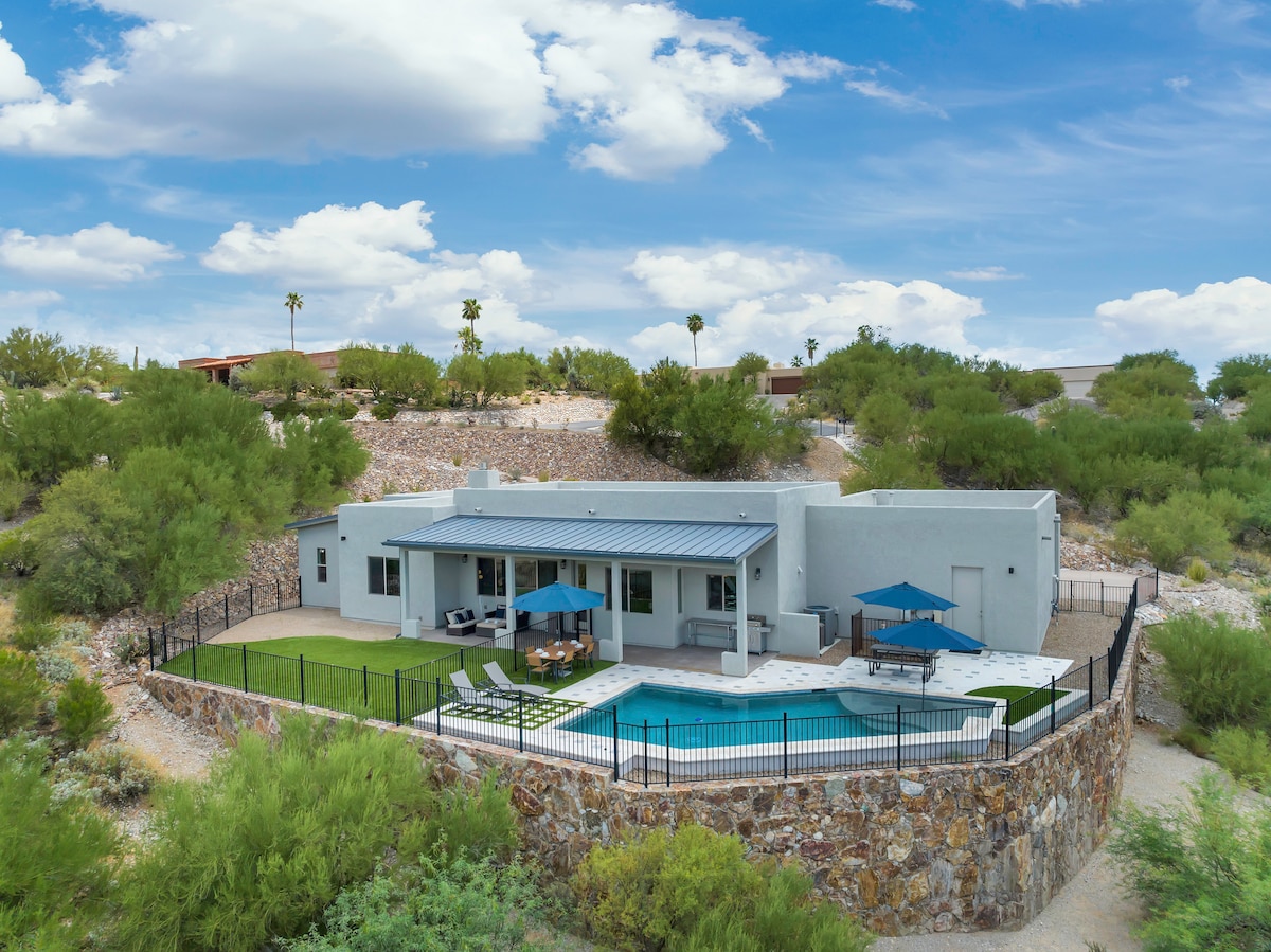 The home is set against a backdrop of lush desert landscaping and features a fenced-in pool area. Blue umbrellas provide shade over lounge chairs next to the pool, with stone accents adding texture to the outdoor space. Mountain views are visible beyond the property.
