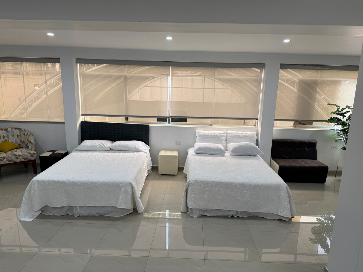 The bright sleeping area is furnished with two spacious beds, each adorned with crisp white linens. Natural light fills the room through large windows, and a cozy armchair complements the minimalist decor. A small side table sits between the beds, adding functionality to the space.