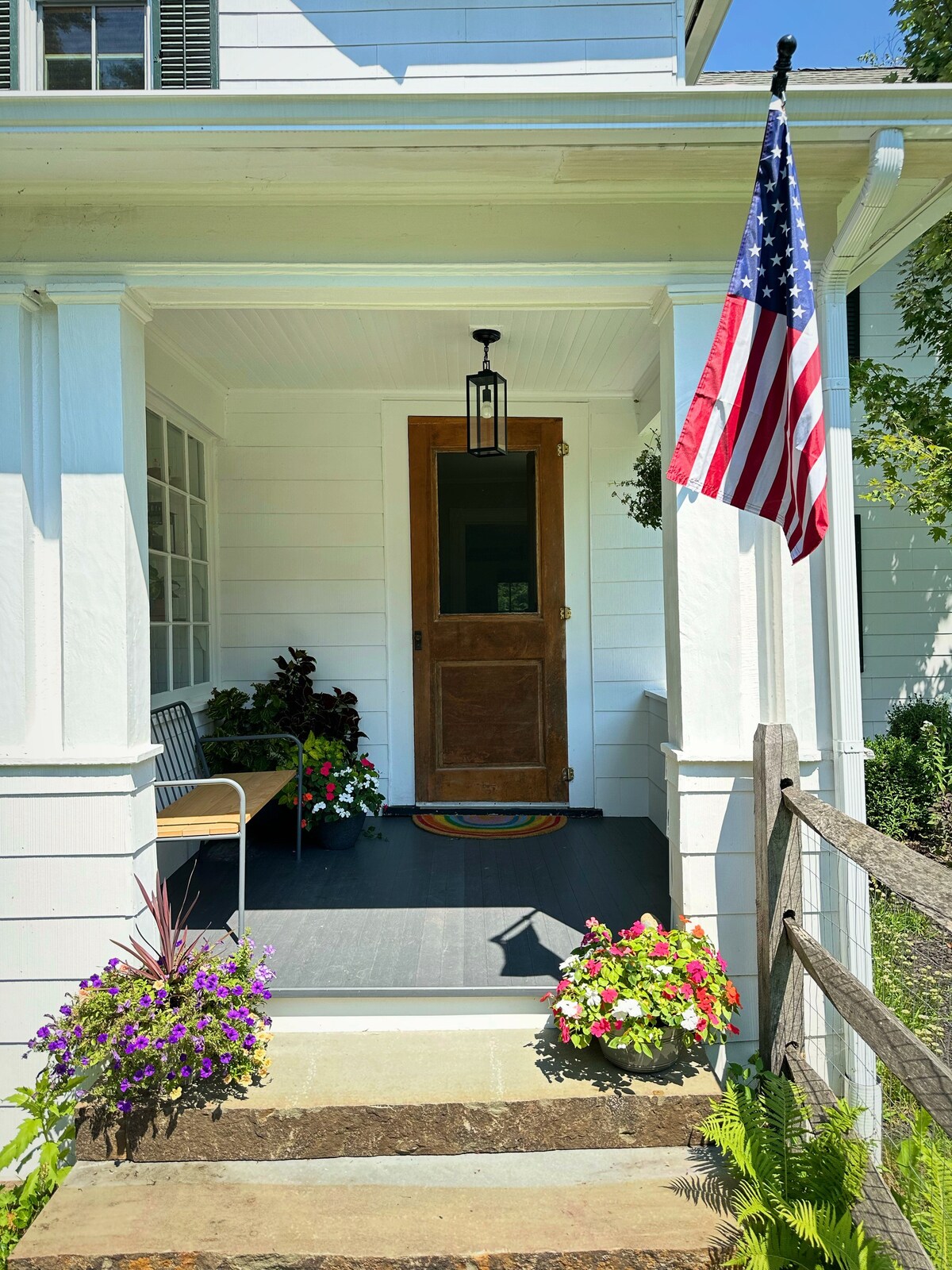 The welcoming entrance features a wooden front door framed by bright flower pots and a flag. A cozy bench sits on the porch, while a colorful rug adds a pop of color. Natural light streams in through large windows, enhancing the charming atmosphere.