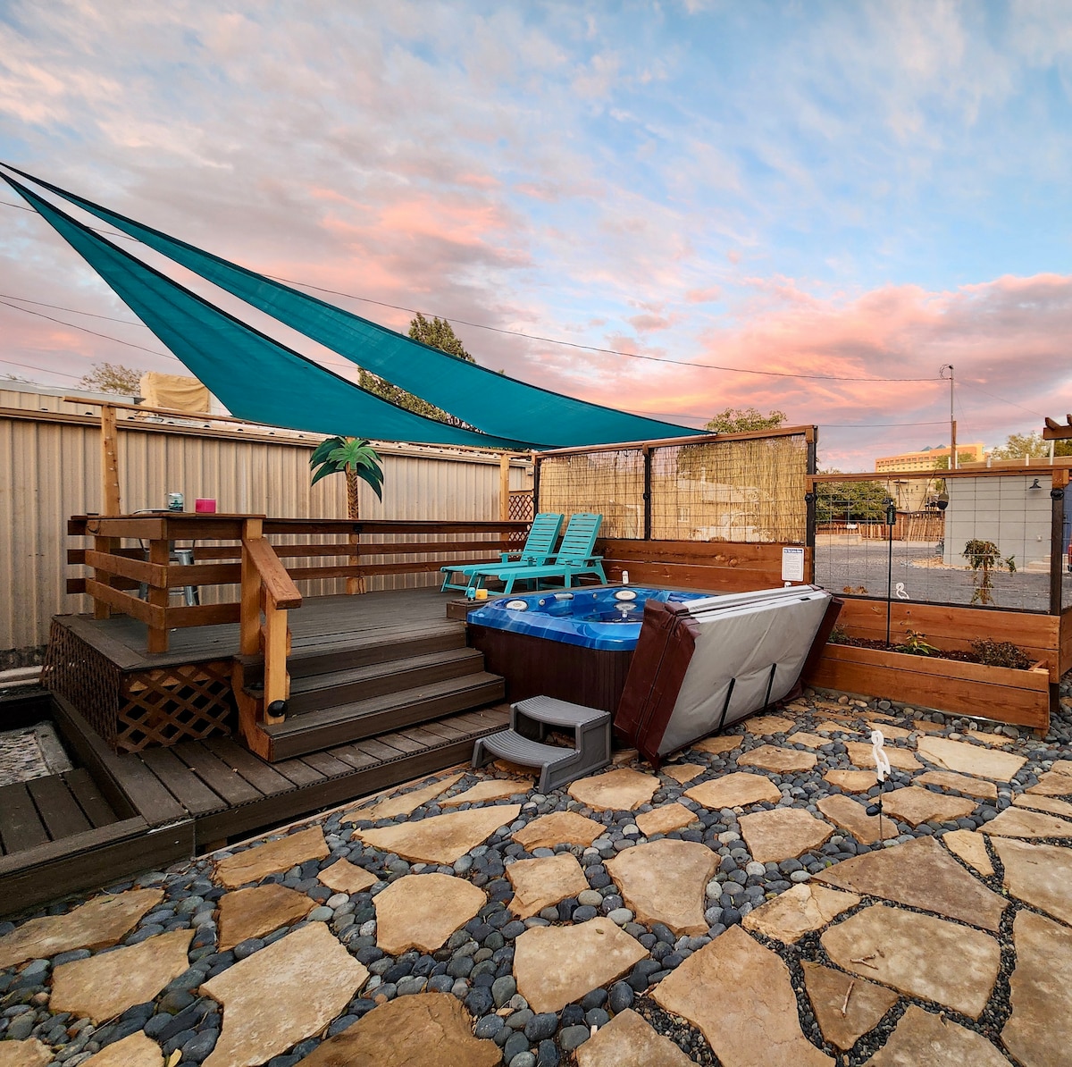 An outdoor area features a private hot tub surrounded by wooden decking. Two blue lounge chairs are positioned nearby under a colorful shade sail. The stone pathway leads to additional landscaped elements, with a visible fence enclosing the space and the sky displaying soft pastel hues.