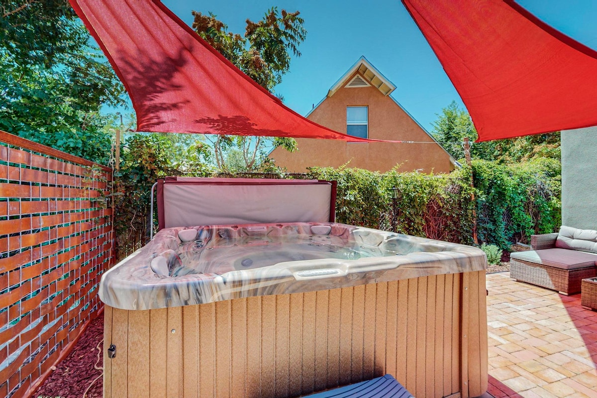 A private hot tub is surrounded by lush greenery and shaded by vibrant red canopies. The space features paver stones and a seating area, creating a relaxing environment in a secluded outdoor setting.